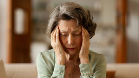 Frau sitzt auf dem Sofa und fasst sich mit beiden Händen an die Stirn